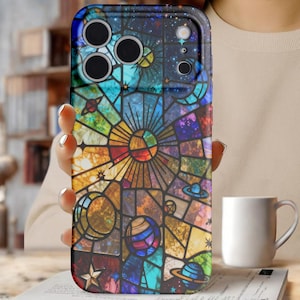 May include: A colorful phone case featuring a stained-glass design with celestial elements. The case showcases planets, stars, and geometric patterns in a vibrant array of colors. The case is held in a person's hand, with a white mug in the background.