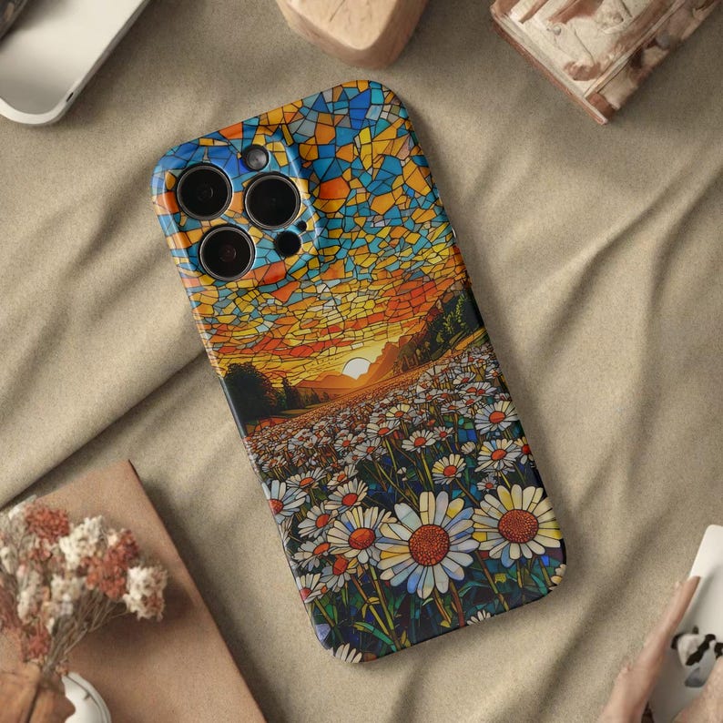 May include: A phone case with a stained-glass effect depicting a sunset over a daisy field. The design features a mosaic sky in shades of orange, yellow, and blue. The foreground shows a field of daisies with white petals and yellow centres.