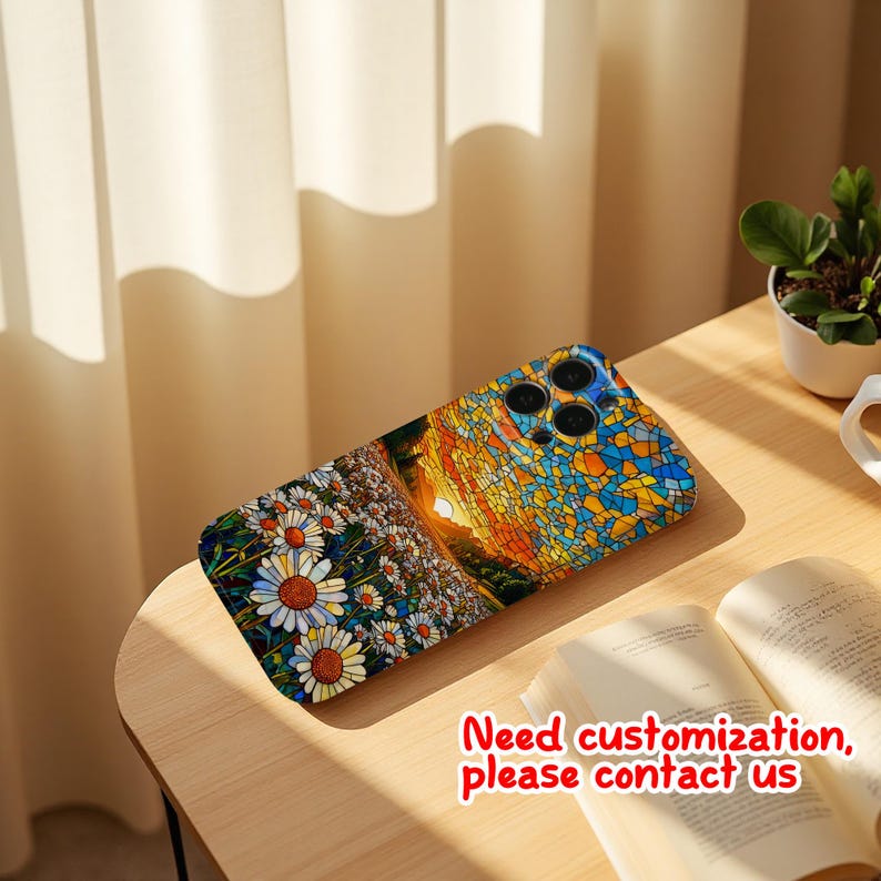 May include: A phone case with a vibrant mosaic design depicting a sunset and a field of daisies. The case has a blue camera module and the text "Need customization, please contact us". It rests on a wooden table.