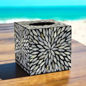 Könnte beinhalten: Ein quadratischer Taschentuchbox-Bezug aus schwarzem Holz mit einem weißen Perlmutt-Einlegemuster. Das Design zeigt ein kreisförmiges Muster aus weißen Blütenblättern, die sich vom Zentrum nach außen erstrecken.