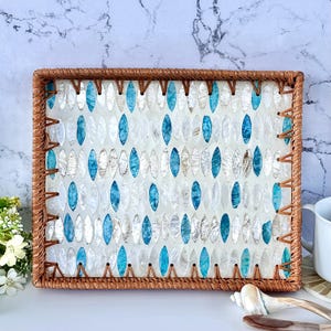 Puede incluir: Bandeja rectangular tejida con una incrustación decorativa de piezas ovaladas blancas, azules e iridiscentes. La bandeja tiene un borde marrón natural y es adecuada para servir o exhibir. El diseño evoca un tema costero o tropical.