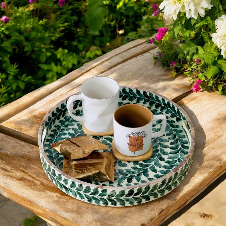 May include: A round wooden tray with a green and white leaf pattern. The tray has two white mugs and a brown mug with a floral design on it. There are also two gold foil-wrapped chocolate bars on the tray.