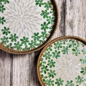 May include: Two round woven trays with a white background and green floral pattern. The trays are made of natural materials and have a rustic look.