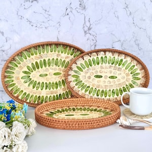May include: Three oval serving trays with woven brown rims. The trays feature a mosaic design of green and off-white pieces. A white mug and decorative flowers are in the background. The trays are a decorative home decor item.