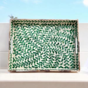 May include: Rectangular decorative tray with a green leaf pattern on a white background. The tray has a wooden frame with a mosaic-like design and integrated handles. The leaf pattern is a repeating design of green leaves.