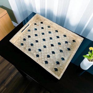 Op de afbeelding: Rechthoekig decoratief dienblad met een crèmekleurig, schelpachtig mozaïekpatroon en donkerblauwe accenten. Het dienblad heeft een lichtgekleurde houten lijst en geïntegreerde handgrepen. Het dienblad staat op een zwart oppervlak.