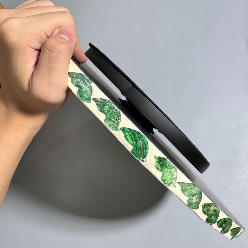 May include: A white round plate with a black backing. The plate has a green leaf design around the edge.