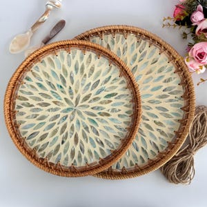 Op de afbeelding: Twee ronde serveerschalen met een geweven rotan rand. Het midden van elke schaal heeft een bloemmotief gemaakt van iriserende, bladvormige stukken in tinten blauw, groen en crème. Een decoratieve lepel en mes zijn zichtbaar.