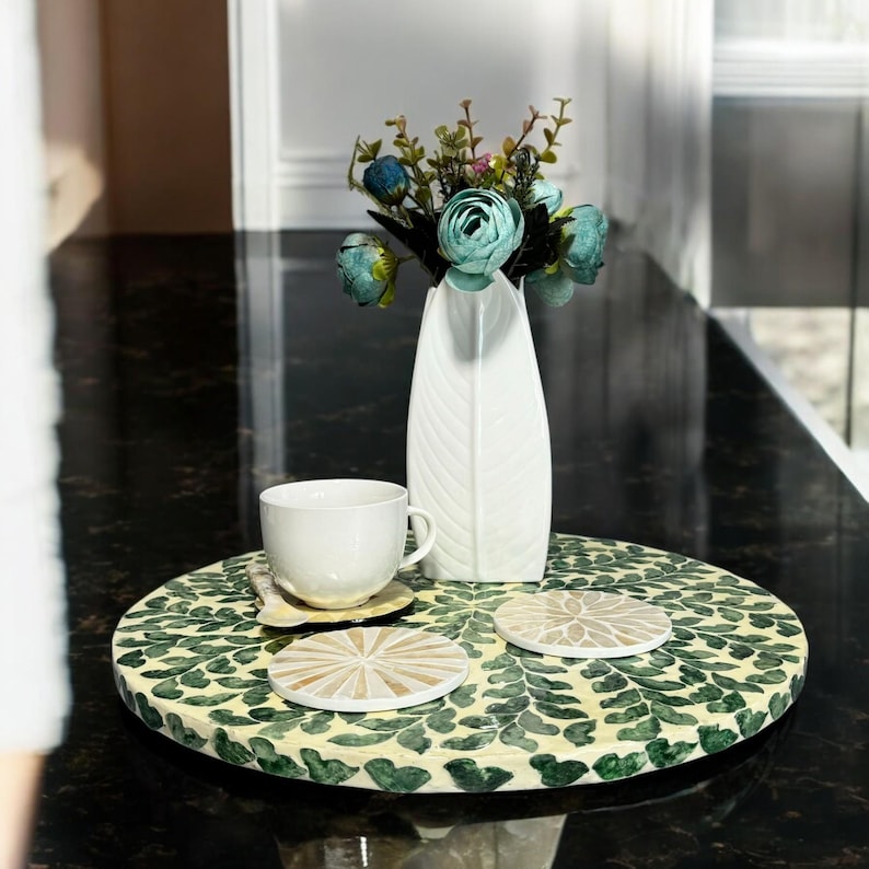 May include: A round, green and white decorative tray with a floral pattern. The tray has three coasters on it, two are white and one is beige. A white vase with a blue flower arrangement is on a black surface behind the tray.