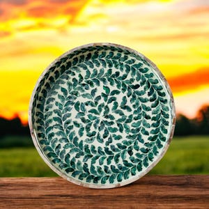 May include: A round decorative bowl with a white background and a green leaf pattern. The bowl is made of mother of pearl and has a shiny finish.