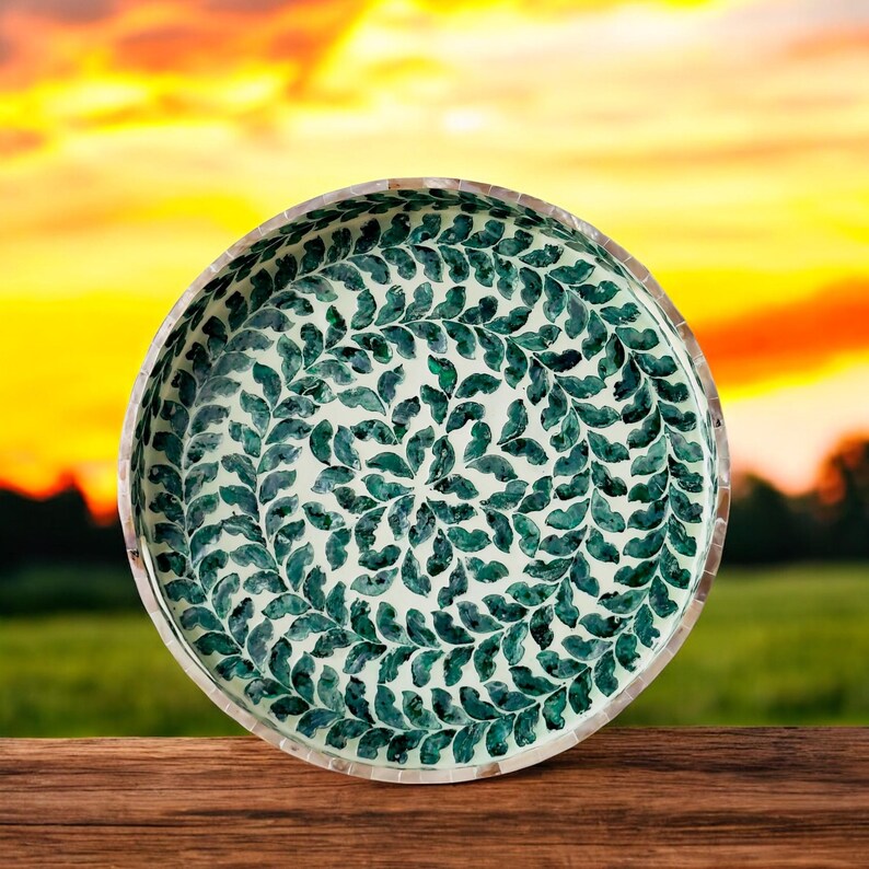 May include: A round decorative bowl with a white background and a green leaf pattern. The bowl is made of mother of pearl and has a shiny finish.