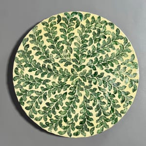 May include: A round ceramic plate with a white background and a green leaf pattern. The leaves are arranged in a radial pattern, creating a sense of movement and symmetry.