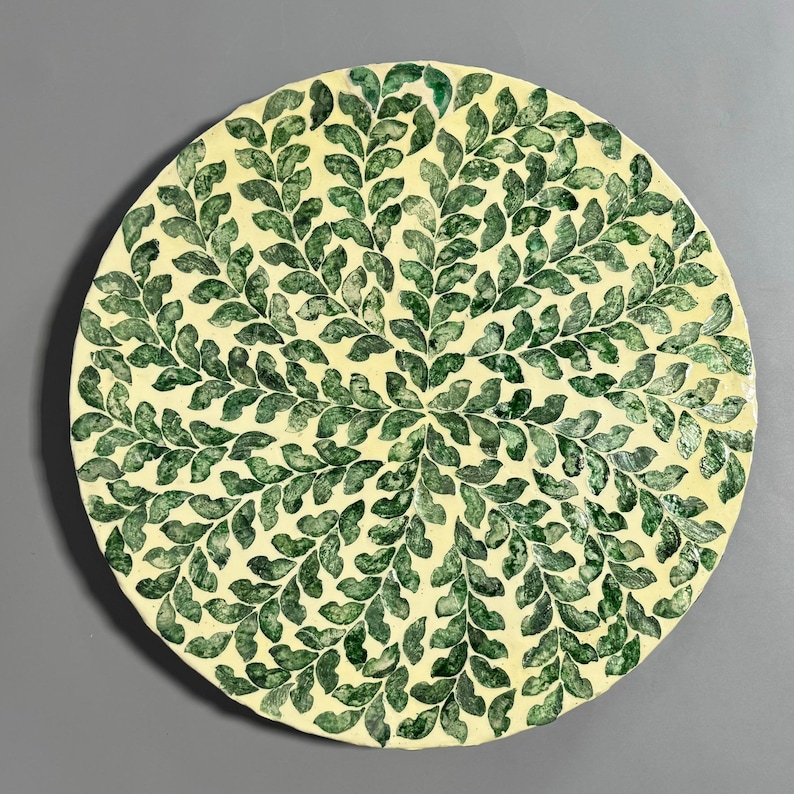 May include: A round ceramic plate with a white background and a green leaf pattern. The leaves are arranged in a radial pattern, creating a sense of movement and symmetry.