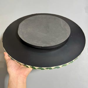 May include: A round, black, decorative tray with a green leaf pattern around the edge. The tray has a black foam base.