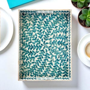 May include: Rectangular wooden serving tray with a cream-colored background and a teal leaf pattern. The tray has a light-colored wooden frame and handles. The leaf design is symmetrical and covers the entire surface of the tray.