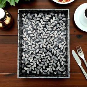 Puede incluir: Una bandeja rectangular con interior negro y un patrón decorativo de hojas hecho de piezas grises y blancas. La bandeja tiene un borde de mosaico y está sobre una superficie de madera.