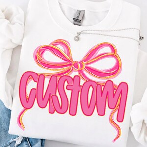 Op de afbeelding: Witte sweatshirt met een roze strik ontwerp en het woord "CUSTOM" in roze letters. Een zilveren ketting met een kleine hanger is over de sweatshirt gedrapeerd. De strik heeft gouden accenten.