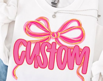 May include: White sweatshirt with a pink bow design and the word "CUSTOM" in pink lettering. A silver necklace with a small pendant is draped over the sweatshirt. The bow has gold accents.