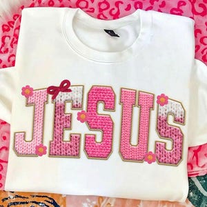 May include: White sweatshirt with the word "JESUS" in pink, knitted-style letters outlined in gold. A small red bow and pink flowers decorate the top of the letters. The sweatshirt is on a pink and white patterned surface.