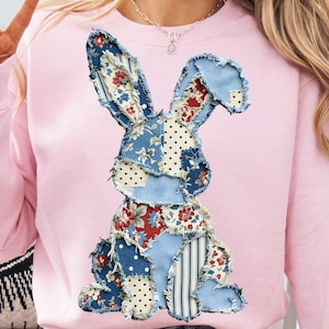 May include: A light pink sweatshirt featuring a patchwork bunny design. The bunny is made of various blue fabrics with floral, polka dot, and striped patterns. The sweatshirt has long sleeves and a crew neck.