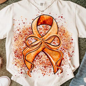 May include: White t-shirt with an orange ribbon design, featuring a leopard print pattern and a bow. The ribbon is set against a backdrop of orange and red paint splatters. The shirt is surrounded by accessories.