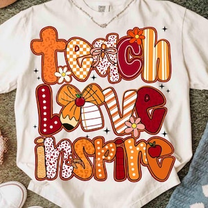 May include: Cream-colored t-shirt with the words "Teach Love Inspire" in a colorful, patterned design. The letters are in shades of orange, red, and white, with floral and apple accents. A casual, short-sleeved tee.