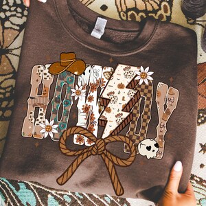 May include: A brown sweatshirt featuring the text "HONEY DAY" in a decorative style. The design incorporates a cowboy hat, flowers, a lightning bolt, and a rope bow. The sweatshirt is held by two hands.