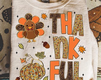 Puede incluir: Una sudadera beige con temática de Acción de Gracias. La palabra "THANKFUL" está impresa en letras naranjas y estampadas. También se muestra un pavo y una calabaza de dibujos animados. El diseño incluye hojas de otoño, bellotas y estrellas.
