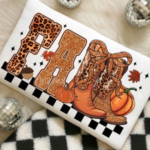 May include: White rectangular pillow with the word "FALL" in leopard print and glittery orange letters. The design includes cowboy boots, pumpkins, and fall leaves. A black and white checkered pattern borders the bottom.