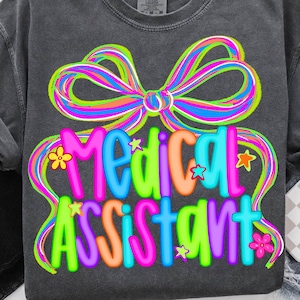 Puede incluir: Camiseta gris oscuro con un lazo colorido y las palabras "MEDICAL ASSISTANT" en letras brillantes. El diseño incluye estrellas y flores. La camiseta se muestra con ropa negra y pantalones cortos de mezclilla.