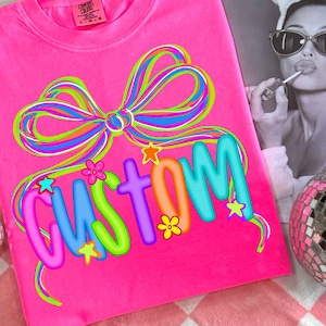 Può includere: T-shirt rosa acceso con un fiocco colorato e la scritta "CUSTOM" in un font giocoso. Il design include stelle e fiori di vari colori. La maglietta è esposta su una superficie a scacchi rosa e bianca.
