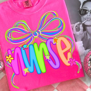 May include: A vivid pink sweatshirt featuring a colourful bow and the word "NURSE" in bright letters. The design incorporates stars and flowers. The shirt rests on a pink and white checked surface, with a disco ball and a monochrome image in the background.