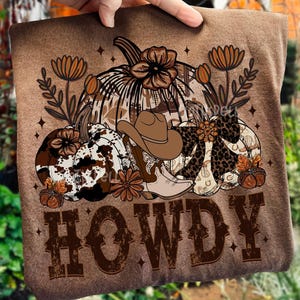 May include: Brown t-shirt featuring a fall-themed design with pumpkins, a cowboy hat, and boots. The design includes floral accents and the word "HOWDY" in a western-style font. The overall color palette is brown and tan.