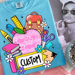 以下が含まれることがあります： 明るいブルーのComfort Colors Tシャツ。学校をテーマにしたグラフィックが特徴です。ピンクのリンゴ、学用品、バックパック、そして「CUSTOM」の文字が黄色い鉛筆型の看板に描かれています。このTシャツはクルーネックで半袖です。