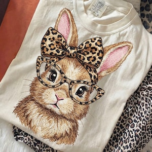 May include: Cream-colored t-shirt featuring a detailed illustration of a bunny wearing glasses and a leopard print bow. The bunny's ears are pink, and the glasses have a cat-eye shape. Leopard print pants are folded next to the shirt.