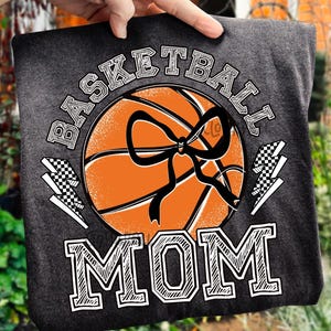 Pode incluir: Camiseta cinza escuro com um design de basquete, um la&ccedil;o preto e as palavras "BASKETBALL MOM" em branco. Dois gr&aacute;ficos de raios ladeiam o basquete. A camiseta &eacute; segurada por uma m&atilde;o.