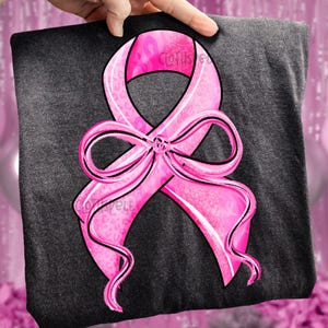 May include: A dark gray t-shirt featuring a large pink ribbon design with a bow. The ribbon has a black outline and a leopard print pattern. The text "Clothspell" is visible on the shirt.