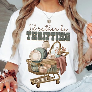 May include: White t-shirt with the text "I'd rather be thrifting" in brown and green. The graphic features a shopping cart filled with dishes, a mirror, and a lamp, suggesting a vintage or antique theme.