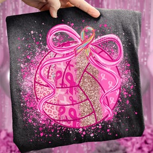 Pode incluir: Camiseta cinza escuro com um design de voleibol rosa, uma fita rosa grande e uma fita multicolorida. O voleibol tem uma seção brilhante e padrões de fita rosa. Salpicos de tinta rosa envolvem o voleibol.