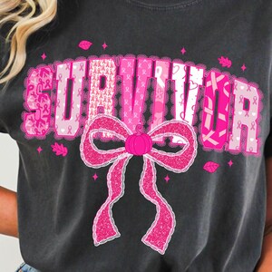 May include: Dark gray t-shirt with the word "SURVIVOR" in pink, featuring a pink ribbon bow and pink ribbon accents. The shirt is a comfortable, casual style, perfect for showing support and raising awareness.