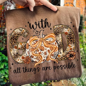 May include: Brown t-shirt with the words "with GOD all things are possible" in a decorative font. The word "GOD" is designed with a leopard print pattern. A pumpkin with a bow is in the center of the design.