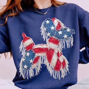 May include: Navy blue sweatshirt featuring a patriotic balloon dog design. The dog is crafted from patches of red, white, and blue fabric, with white star accents and fringed edges. A casual, comfortable top for everyday wear.
