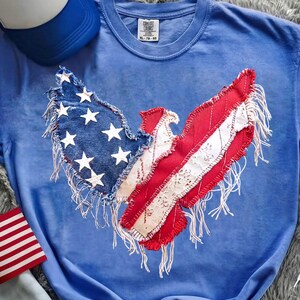 May include: A blue Comfort Colors t-shirt featuring a patriotic eagle design. The eagle's wings are crafted from denim with white stars and red and white striped fabric, fringed edges. A blue baseball cap and a small American flag are visible.
