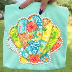 May include: A light teal sweatshirt featuring a seashell design. The seashell is made of colorful patchwork with tropical flowers, seahorses, and pineapples. The seashell is outlined with pearls and gold trim.