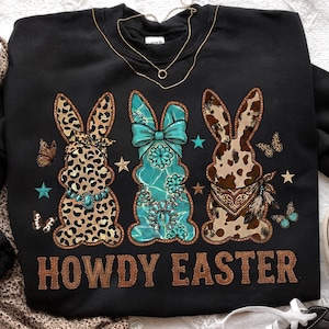 May include: Black sweatshirt with a "Howdy Easter" graphic. The graphic features three bunny figures with leopard, turquoise, and cowhide patterns. Butterflies and stars accent the design. A gold necklace is draped on the sweatshirt.