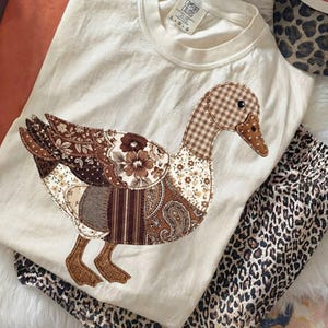 Könnte beinhalten: Cremefarbenes T-Shirt mit einem Patchwork-Enten-Design in Brauntönen. Die Ente weist verschiedene Muster auf, darunter Blumen, Paisley und Vichy. Eine Hose mit Leopardenmuster ist sichtbar. Das Shirt hat ein Comfort Colors-Etikett.