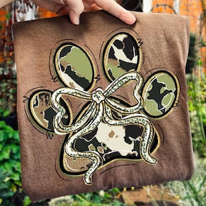 K&ouml;nnte beinhalten: Braunes T-Shirt mit einem Pfotenabdruck-Design und einer Schleife. Der Pfotenabdruck besteht aus vier ovalen Formen und einer zentralen Schleife, alle mit einem Tarnmuster. Das Design ist in Gold und Schwarz umrandet.