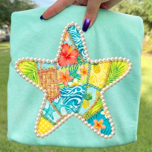May include: A light turquoise shirt featuring a large, star-shaped appliqué. The star is composed of various fabric patches with tropical patterns, including flowers, pineapples, and leaves. The star is outlined with faux pearls.