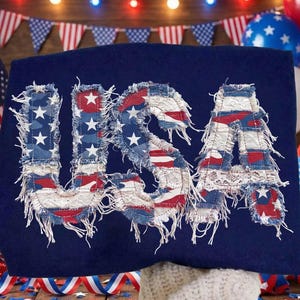 Könnte beinhalten: Ein marineblaues T-Shirt mit dem Wort "USA" in einem patriotischen Design. Die Buchstaben zeigen einen Patchwork aus roten, weißen und blauen Stoffen, einschließlich Sterne und Streifen, mit ausgefransten Kanten. Das T-Shirt ist für einen patriotischen Feiertag dekoriert.