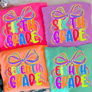 May include: Four colorful t-shirts with grade level text. The shirts are pink, purple, orange, and teal. Each shirt has a large bow design with the words "Fifth Grade", "Sixth Grade", "Seventh Grade", and "Eighth Grade" in a rainbow font.